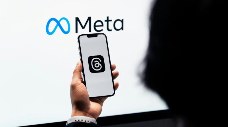 Mãos segurando um smartphone com o logotipo da Meta Threads na tela e a marca Meta ao fundo.