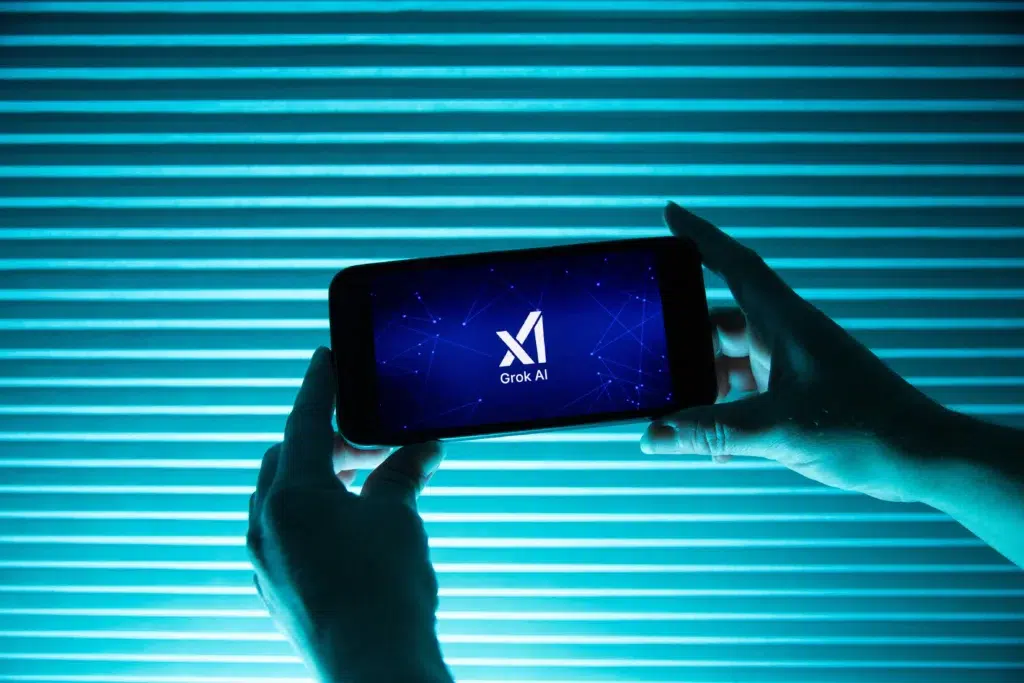 Pessoa segura telefone exibindo logo da inteligência artificial Grok, do Elon Musk, com fundo azul; polêmica sobre discurso antissemita em 2025 na X.