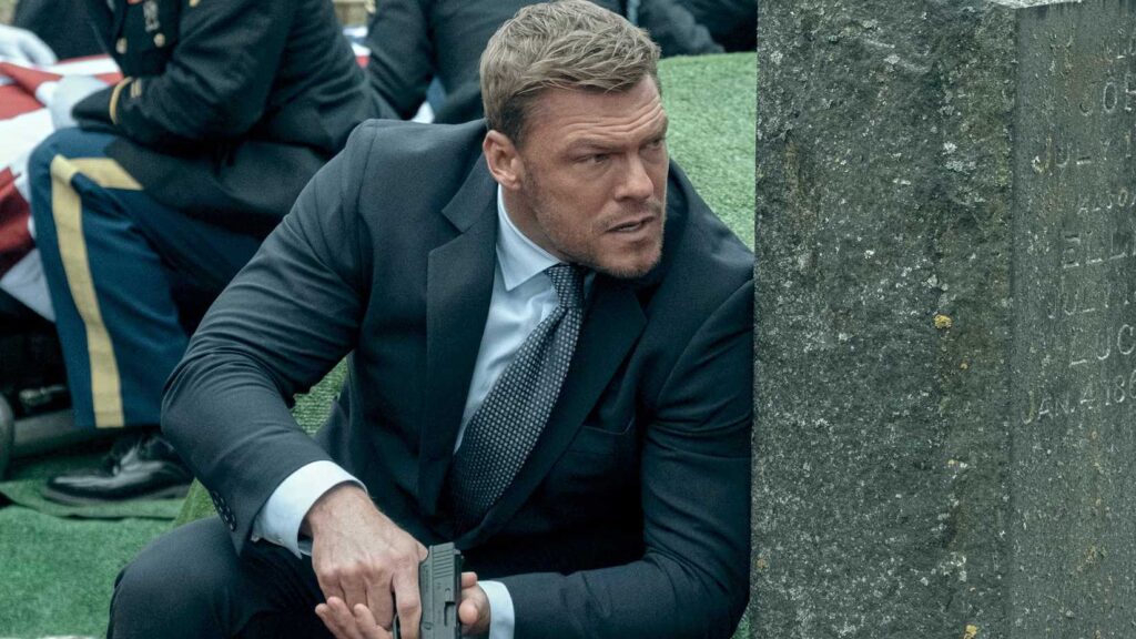 Alan Ritchson interpreta Jack Reacher, se protegendo atrás de um túmulo.